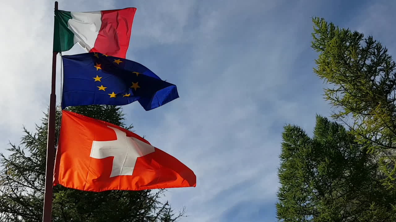 Bandiere di Italia, Europa e Svizzera che sventolano insieme al vento sulle Alpi italiane.