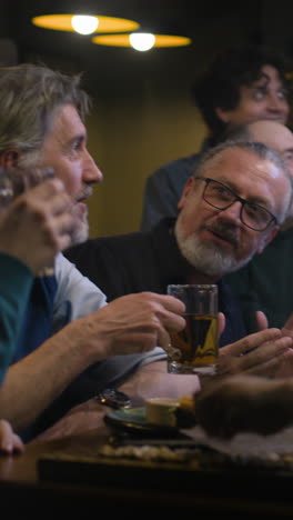 Vertical-shot-of-friends-rejoice-and-celebrate-when-football-team-scores-goal-and-wins-world-league-championship.-Happy-sports-fans-shout-and-drink-beer-watching-soccer-match-live-in-sports-bar-or-pub-at-night.