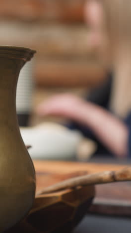 clay jug and plate with spoon on table in yard near wooden house. young woman writes sitting at table near handmade dishes on blurred background