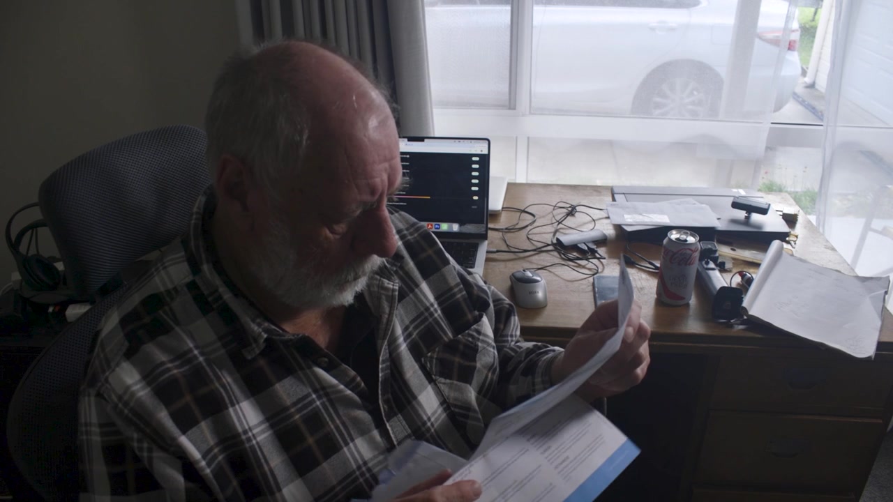 An older man sits at a home desk, opening and reading a letter. Nearby, a laptop and a drink can be seen. Suitable for depicting everyday life or personal finance themes.