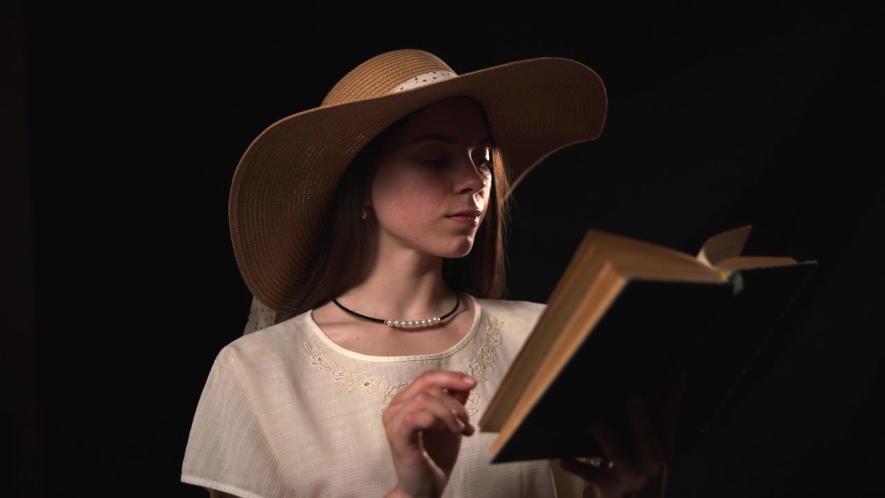 Una giovane donna con un cappello a tesa larga legge un libro aperto su uno sfondo scuro, delicatamente illuminata frontalmente con il libro visibile in primo piano e il suo viso.