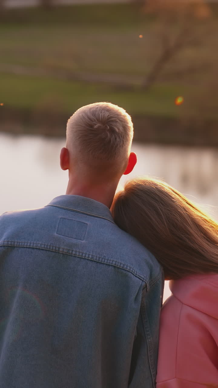 Young couple near the river. Guy and his girlfriend standing together and looking at sunset among beautiful nature. Rear view. Vertical video