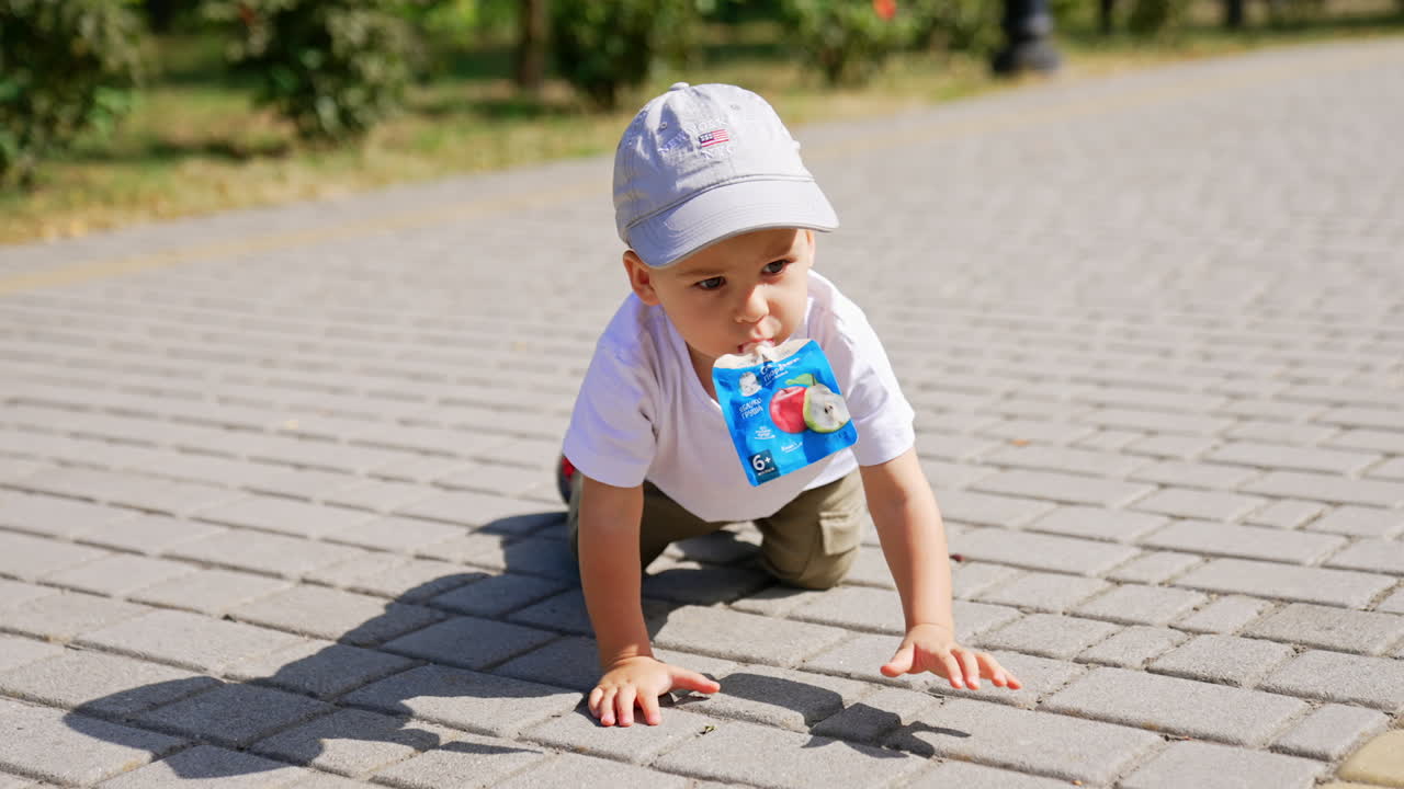 Kid in a cap holding a doy pack in his mouth crawls by the paved road. Child outdoors on sunny summer day.