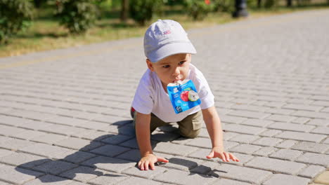 kid in a cap holding a doy pack in his mouth crawls by the paved road. child outdoors on sunny summer day.