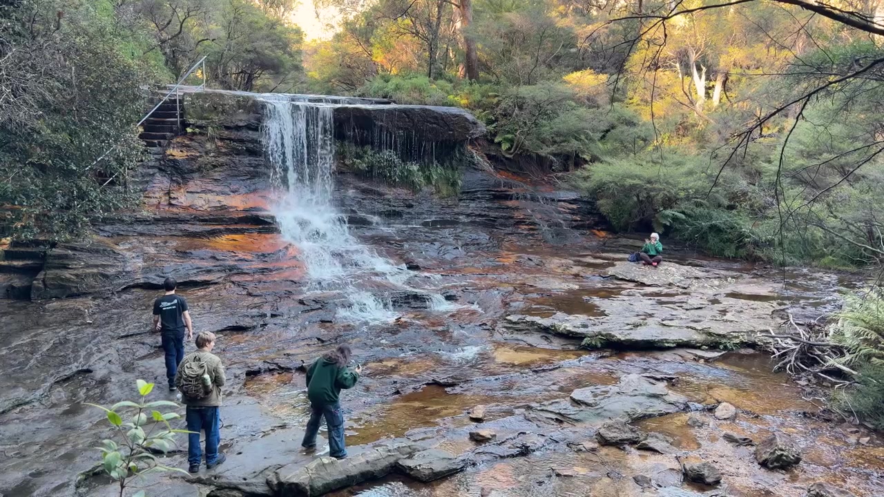 Turister fotograferar ett naturskönt vattenfall över klippig terräng i Wentworth Falls, Blue Mountains, Australien.