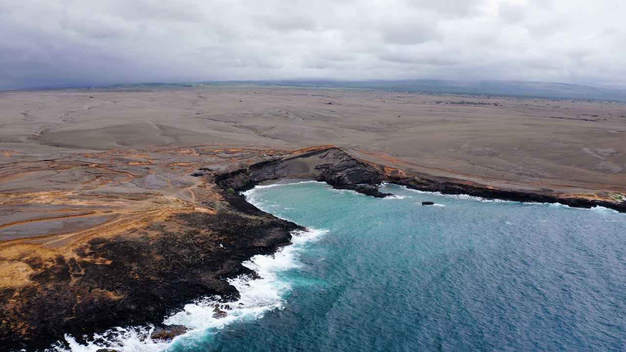 Dramatic volcanic coastline with dark rock formations and waves crashing against the shore. The expansive landscape stretches inland beneath a cloudy sky.