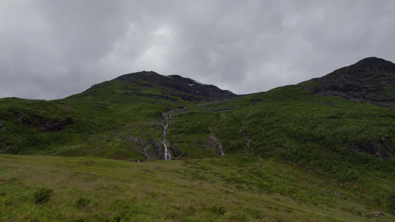 Tynne fossefall slynger seg ned en gresskledd grønn fjellside under tunge grå skyer i Norangsdalen, Sunnmøre, Norge