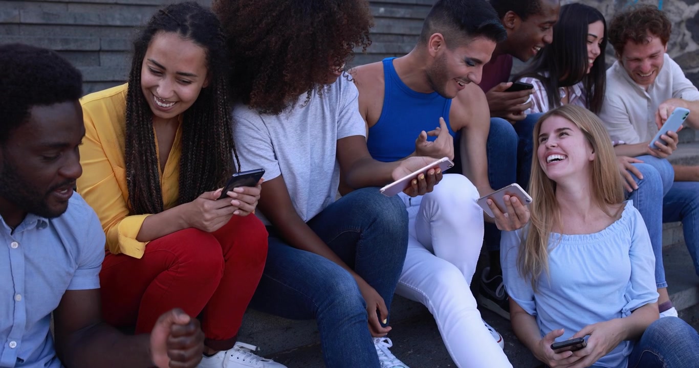 Gruppo eterogeneo di giovani amici che ridono mentre usano gli smartphone all'aperto