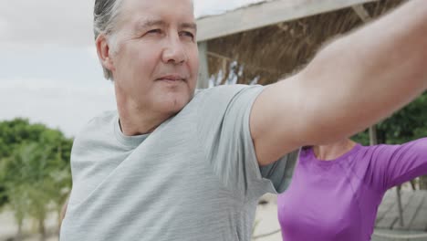 Happy-senior-caucasian-couple-practicing-yoga-standing-on-beach-sun-deck,-in-slow-motion