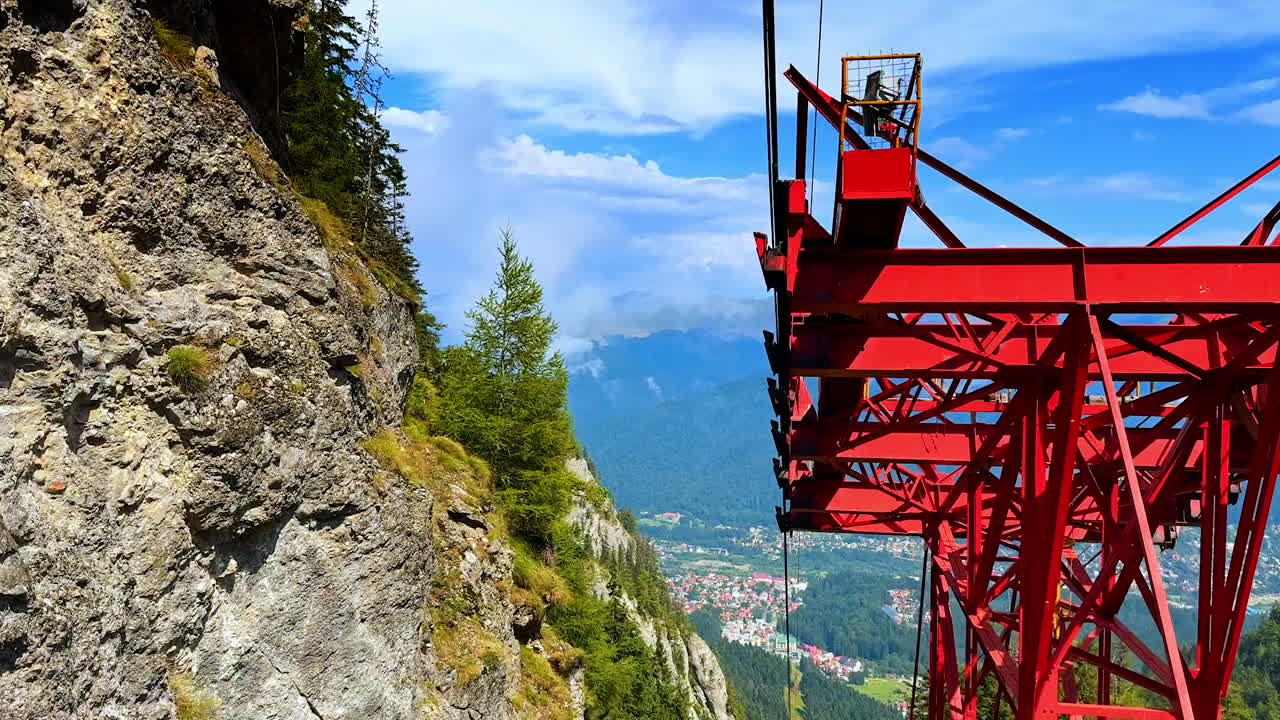 View from inside a red cable car gondola. The metal structure frames a steep rocky cliff and the distant valley below