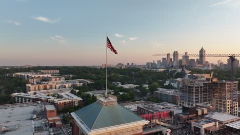 Bandera-Americana-Ondeando-En-La-Cima-De-La-Torre-Del-Mercado-De-La-Ciudad-De-Ponce-Con-La-Vista-Del-Paisaje-Urbano-De-Atlanta-Al-Fondo-Durante-La-Puesta-De-Sol,-Georgia,-EE.UU.