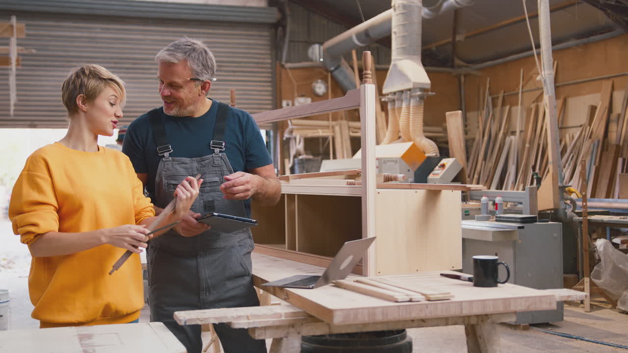 Female Apprentice Learning From Mature Male Carpenter With Digital Tablet In Furniture Workshop