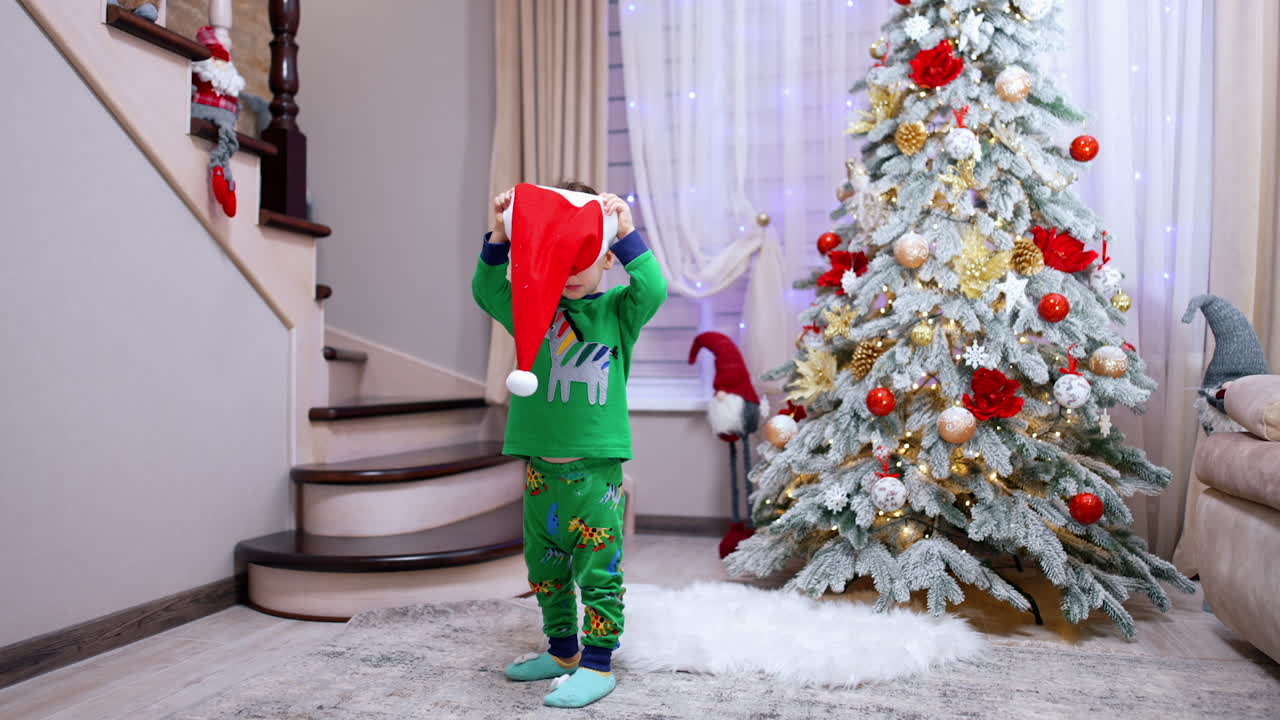 Kid wearing green pajama stands in the room with beautiful Christmas tree. Baby boy puts red Santa cap on.