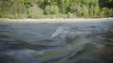close-up, surface-level shot capturing the textured flow of the arachthos river as it ripples toward the viewer, with a tree-lined bank and gravel shore in the background.