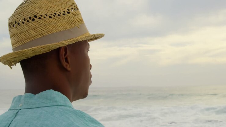 Vista laterale di un uomo afroamericano con un cappello in piedi sulla spiaggia 4k