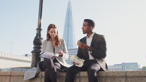 Two-millennial-colleagues-take-a-break-on-the-embankment-eating-and-talking-sitting-on-the-Thames-wall-by-the-river-near-London-Bridge,-low-angle,-close-up