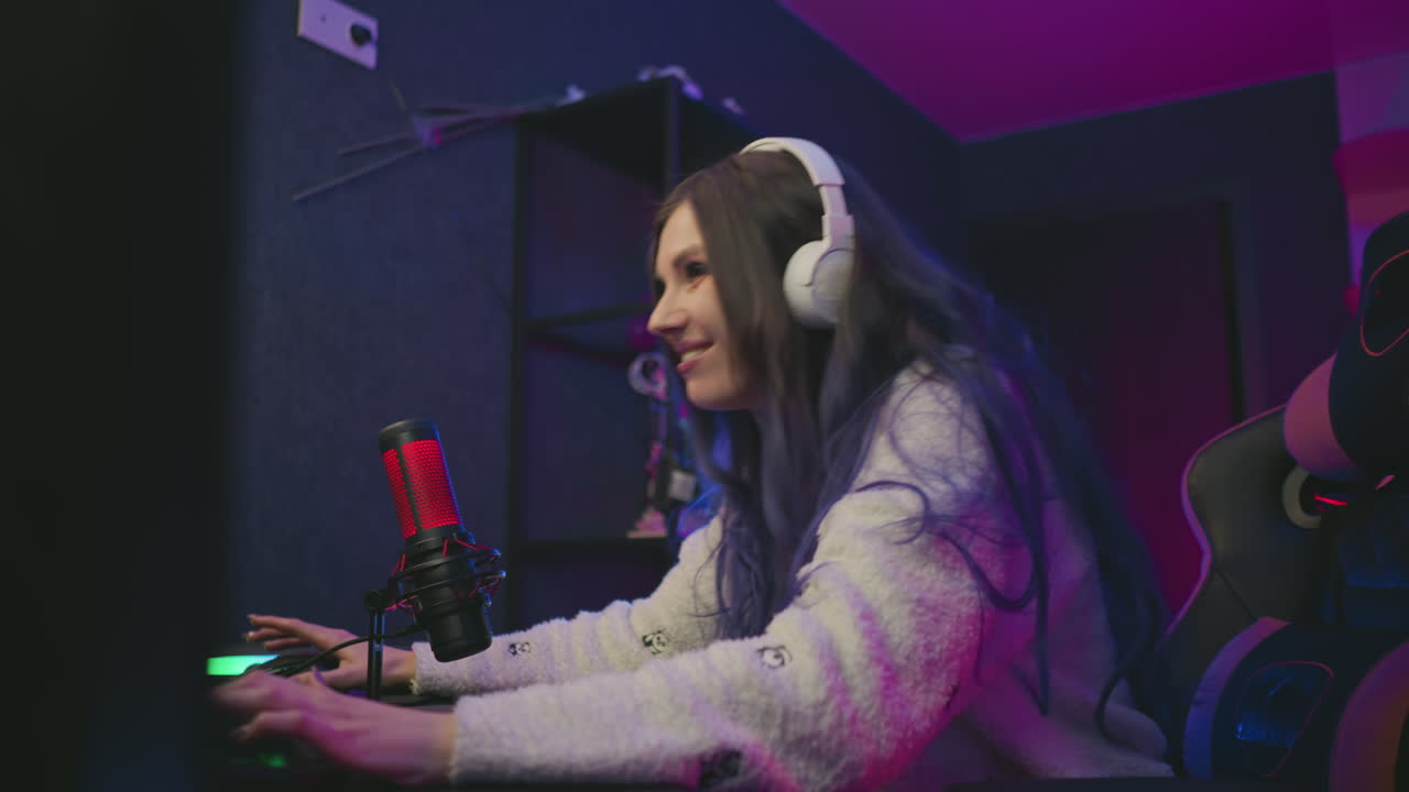 side view of young gamer with long hair and headphones seated in gaming chair playing computer game, showing excitement after winning, surrounded by colorful studio lights and condenser microphone