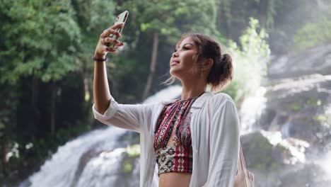 woman with her hair tied back stands beside a waterfall, capturing the moment with her smartphone