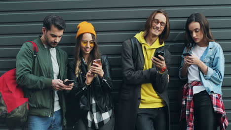 grupo de amigos caucásicos apoyados en la pared mientras hablan y escriben en sus smartphones en la calle