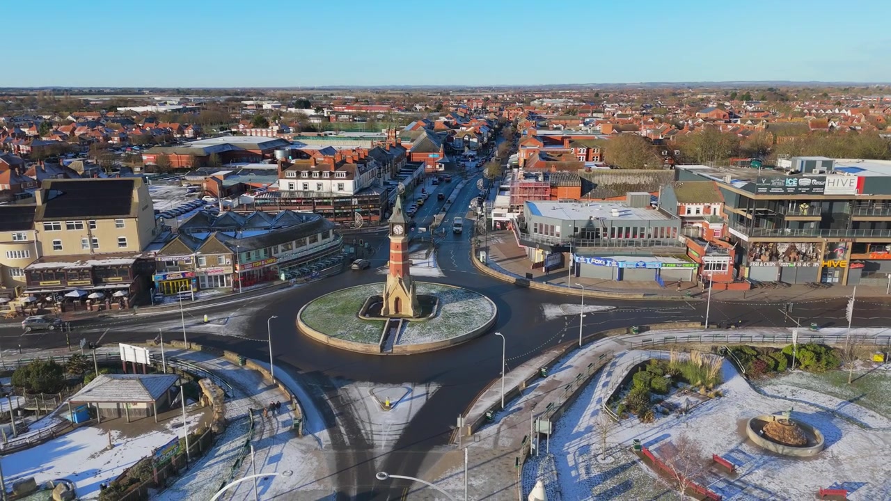 Luftfoto af et bytorv med rundkørsel og tårn. Sne dækker gader og bygninger under en klar himmel. Køretøjer navigerer roligt i den cirkulære krydsning.