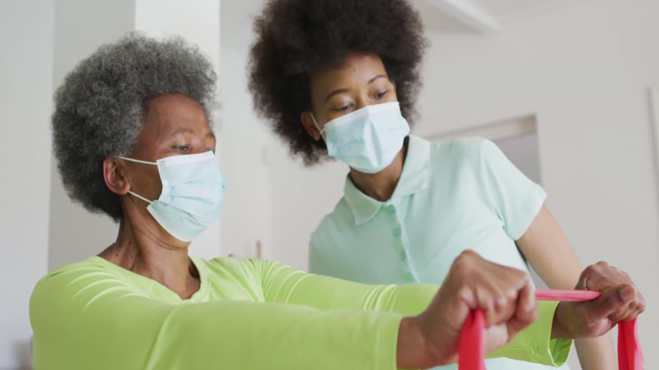 African american female physiotherapist wearing face mask helping senior female patient exercise
