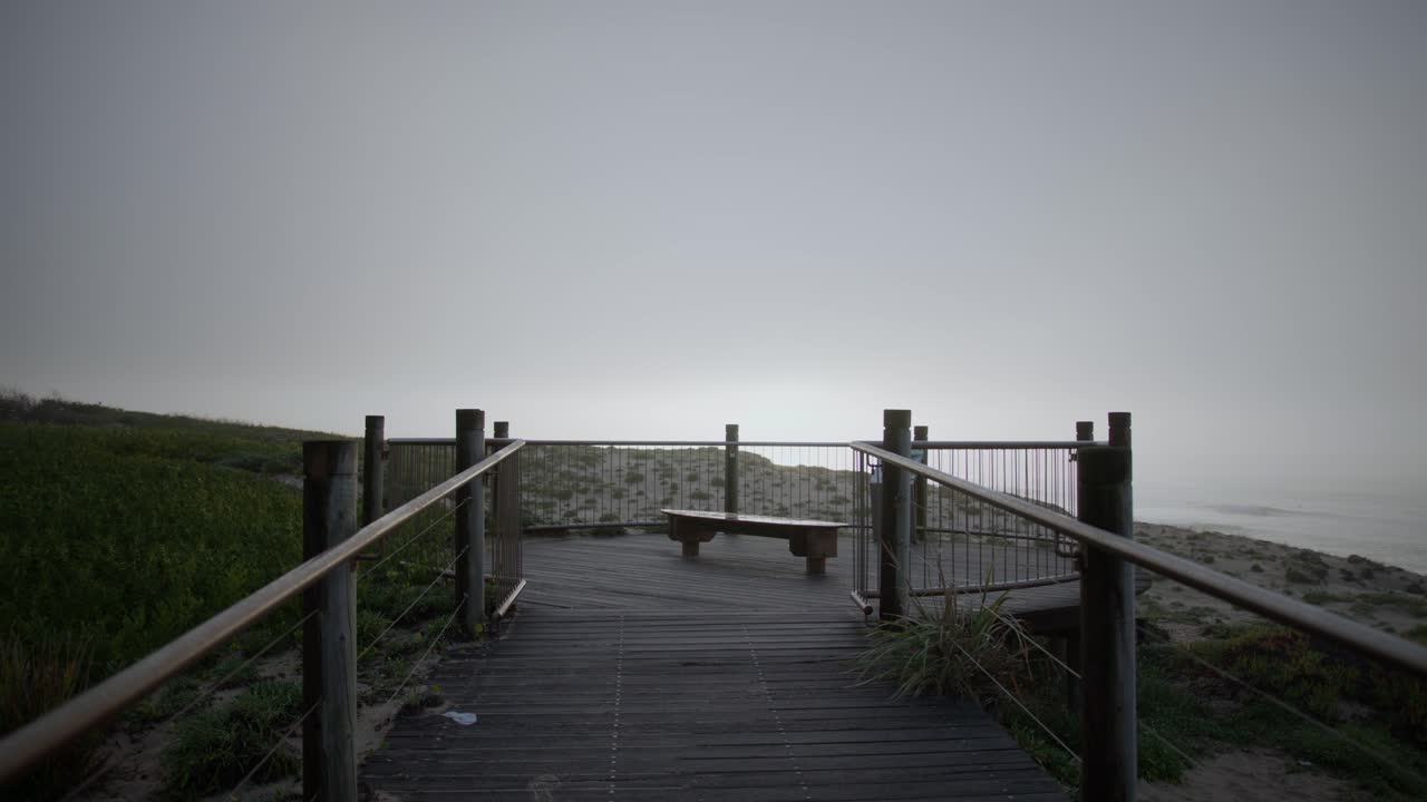 Morbida nebbia mattutina avvolge le dune costiere mentre la luce del sole filtra sull'oceano, un surfista solitario visibile in lontananza, evocando isolamento, calma e tranquilla introspezione.