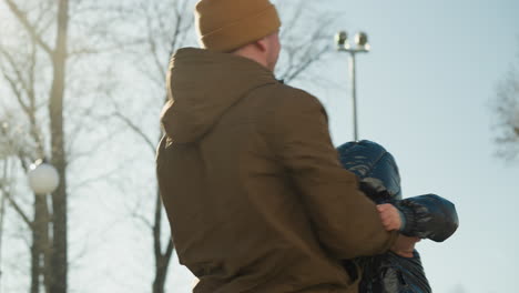 a father playfully spins his son around gently in an outdoor park, lifting him up and down with joy, with trees in the background