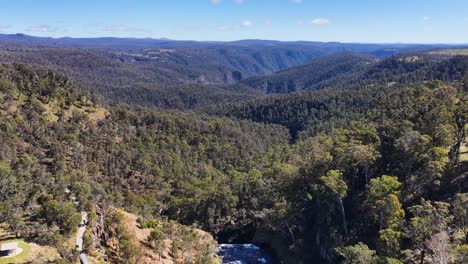 drone footage glides above ebor gorge near tamworth, revealing winding river, rugged cliffs, dense eucalyptus forest, and distant mountain ranges under bright daylight