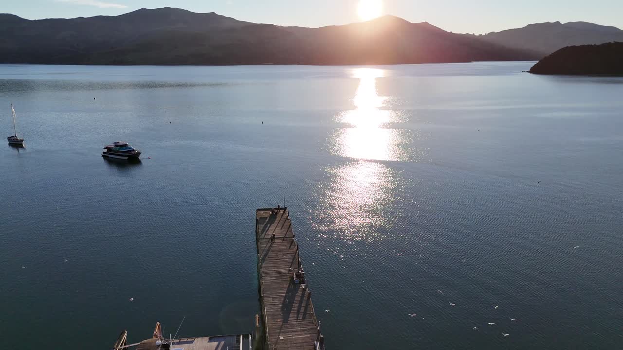 Aerial view of Akaroa harbor at sunset, capturing boats, calm waters, and sun reflections