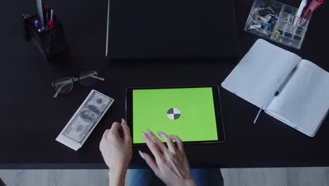 woman's hands use digital tablet on desktop with laptop, money dollars, glasses and other office supplies, closeup, top view. business concept