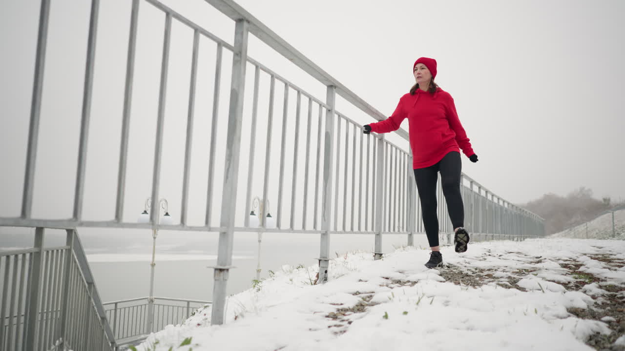 mujer haciendo ejercicio al aire libre durante el invierno, sosteniendo una barandilla de hierro para el equilibrio mientras estira su pierna hacia adelante y hacia atrás en el suelo nevado, atmósfera brumosa en el fondo, postes de luz y río lejano
