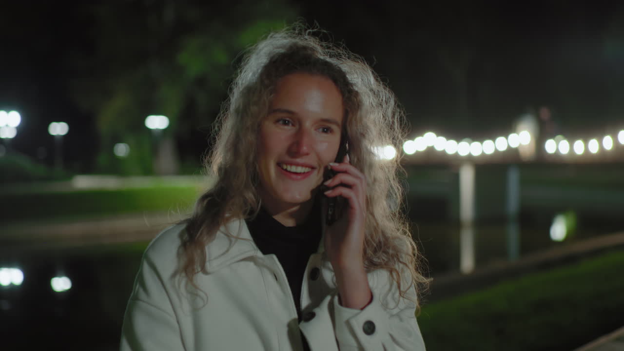Joyful white lady with curly hair sits outside at night on phone call, hair swaying gently under warm lights, blurred reflection behind over calm water