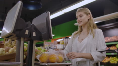 a blonde girl in a supermarket weighs oranges on an electronic scale pressing the display standing with a basket in her hands