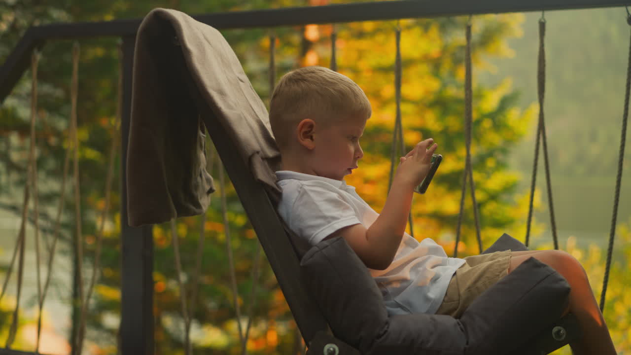 Serious boy holds cellphone sitting alone on balcony at sunset. Focused toddler kid plays video games enjoying atmospheric location with incredible view in background