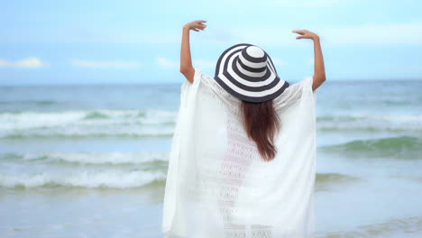 a young woman in beach clothes and a big hat walks on the sandy beach with arms outstretched on the beach and enjoys the view of the sea