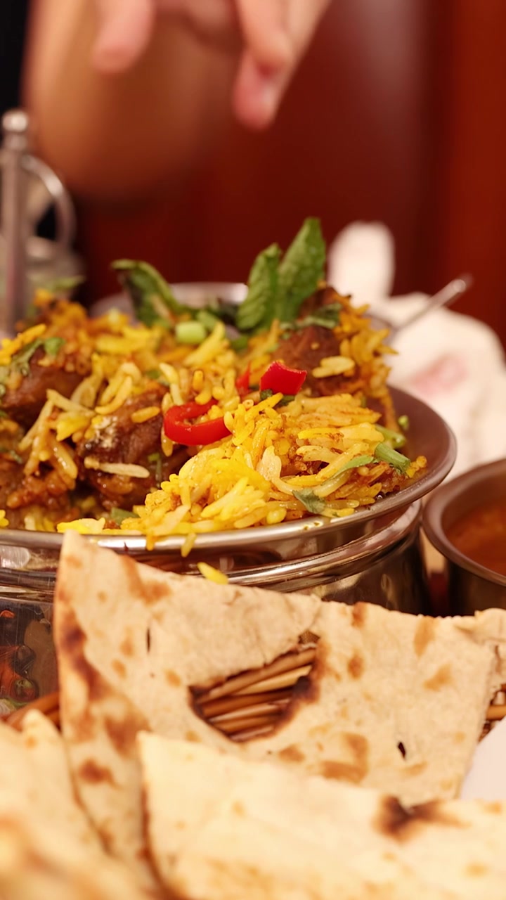 Primer plano de manos sirviendo arroz biryani amarillo de un cuenco de metal junto a naan y una olla de barro