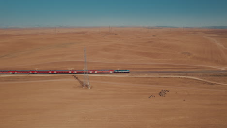 aerial shot of a red passenger train in a remote and desert area, near large electricity poles, dry land without crops, on the ground there is a female shepherd with sheeps