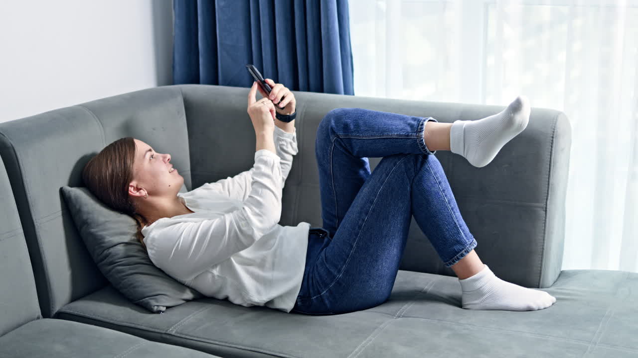 Donna felice e positiva a casa sul divano che naviga e scorre un'app di social media sul cellulare/smartphone. Donna che lavora da casa in remoto.