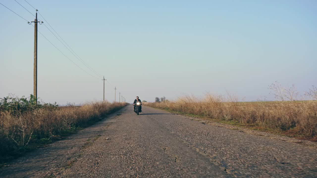 선글라스와 가죽 재 ⁇ 을 입은 세련된 시원한 젊은이가  ⁇ 은 날 도로에서 오토바이를 타고 다가오고 있습니다. 그리고 그는 카메라의 시점을 뛰어넘어 앞으로 나아갑니다. 4k로 촬영되었습니다.