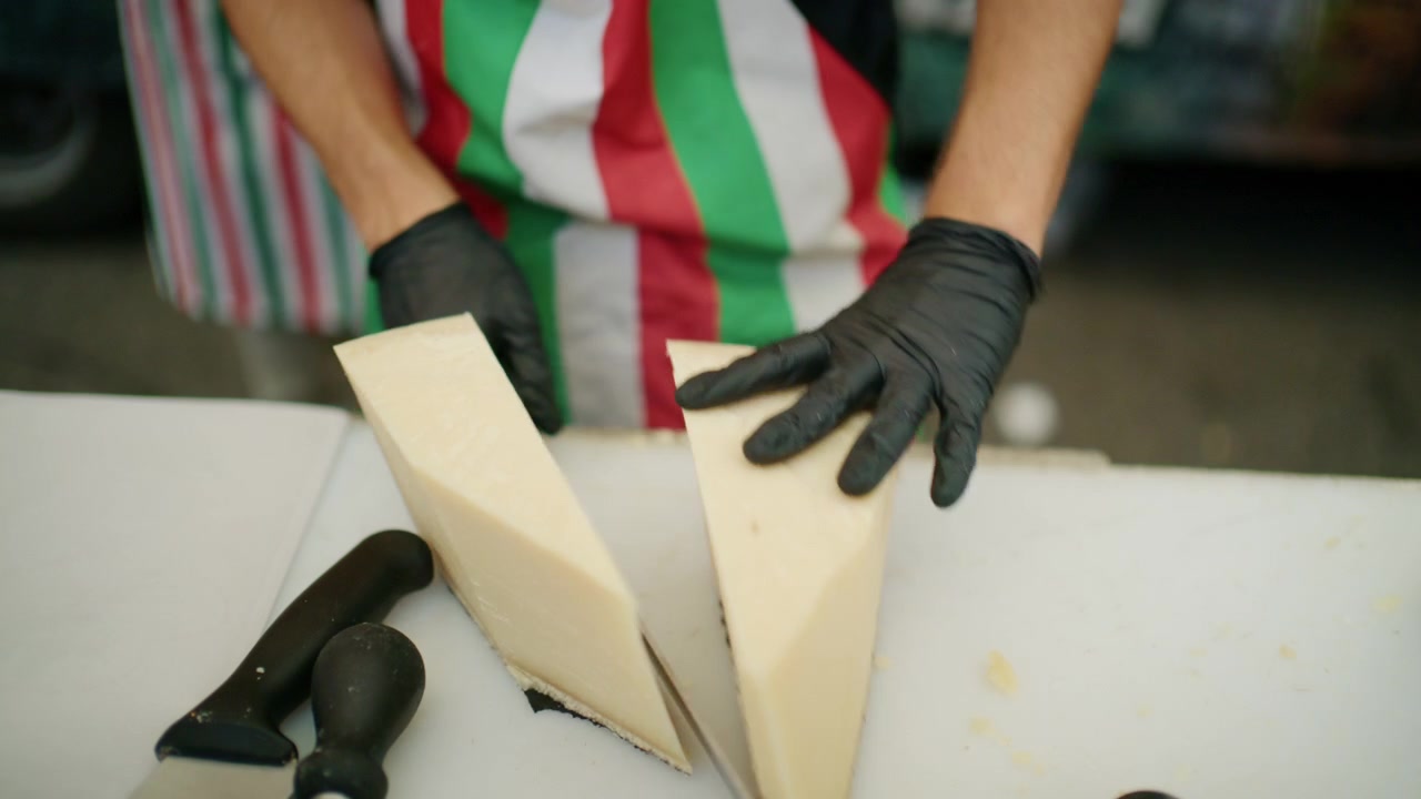 Chef con grembiule dai colori della bandiera italiana che affetta un grande spicchio di formaggio su un tagliere bianco.