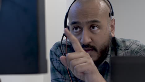 bald uk asian male talking to colleague wearing headset microphone viewed behind monitor