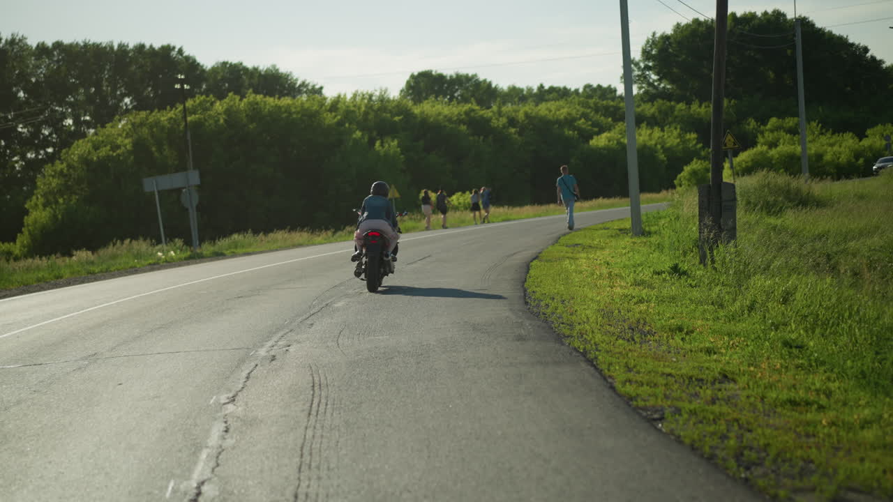 dos mujeres montando una bicicleta eléctrica alrededor de una curva, con una vista de personas caminando por el lado de la carretera y un coche que se acerca desde la dirección opuesta, la carretera está rodeada de vegetación