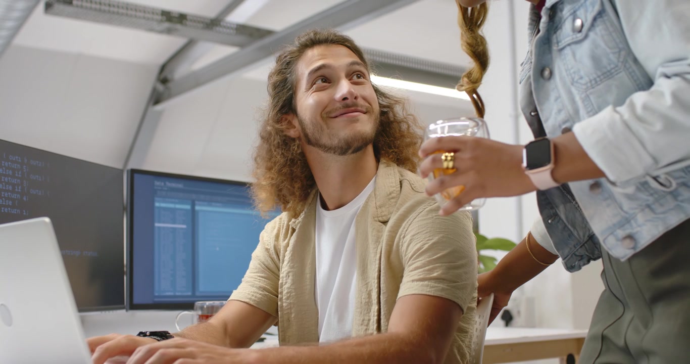 Coworkers, woman entering offering glass mug, causing man pausing work on laptop at desk smiling