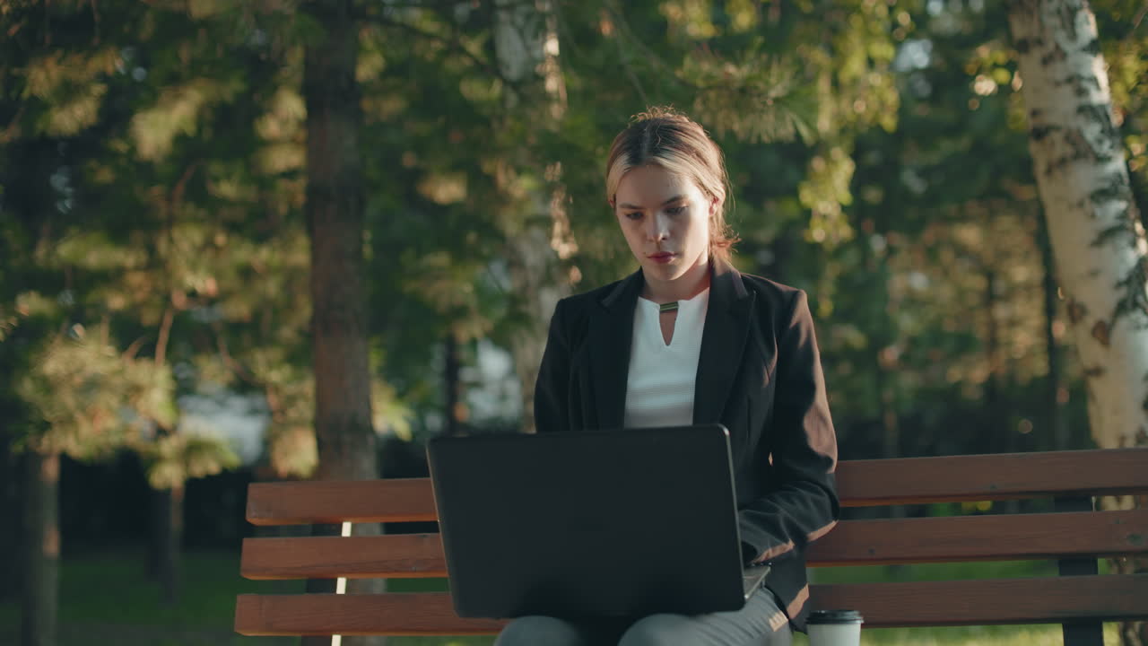 Una dattilografa concentrata si ferma per sorseggiare una bevanda mentre lavora all'aperto con un laptop su una panchina di legno, con la luce del sole che le illumina il viso e gli alberi che la circondano in un tranquillo parco.
