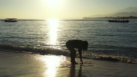 silhouette of girl washing her feet on warm clear water of sea at sunset moments over lagoon with boats, bali