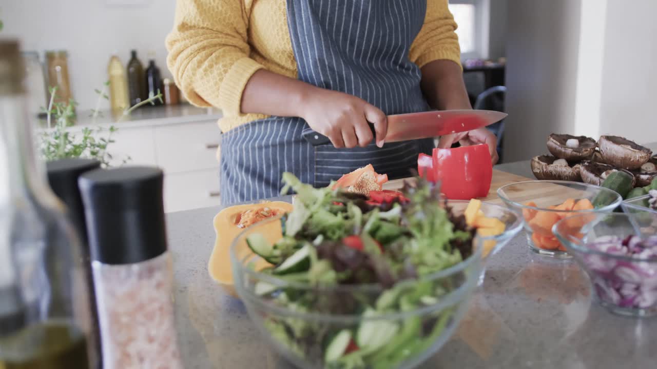 sección media de una mujer afroamericana en delantal cortando verduras en la cocina, cámara lenta