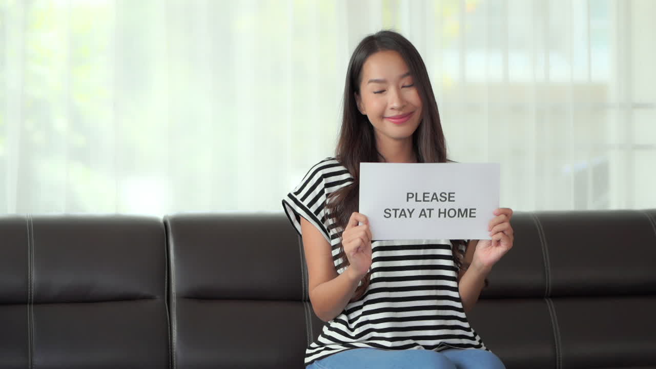 A pretty young Asian woman sitting on a couch holds up a sign "Please Stay at Home." copy space