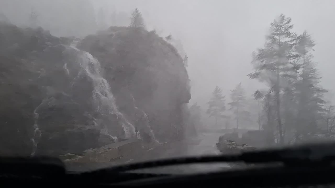 Car drives in stormy conditions with water pouring on windshield. Wipers move fast as visibility drops while passing wet road near powerful waterfall