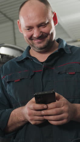 inventor en uniforme azul sonriendo mientras opera su teléfono en un taller automotriz, haciendo una pausa para comprobar algo antes de centrarse de nuevo en el dispositivo, con herramientas mecánicas en el fondo