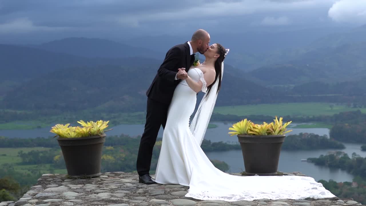 The groom giving a long, romantic kiss to the bride on their wedding day. Background: cloudy mountains, dreamy scenery.